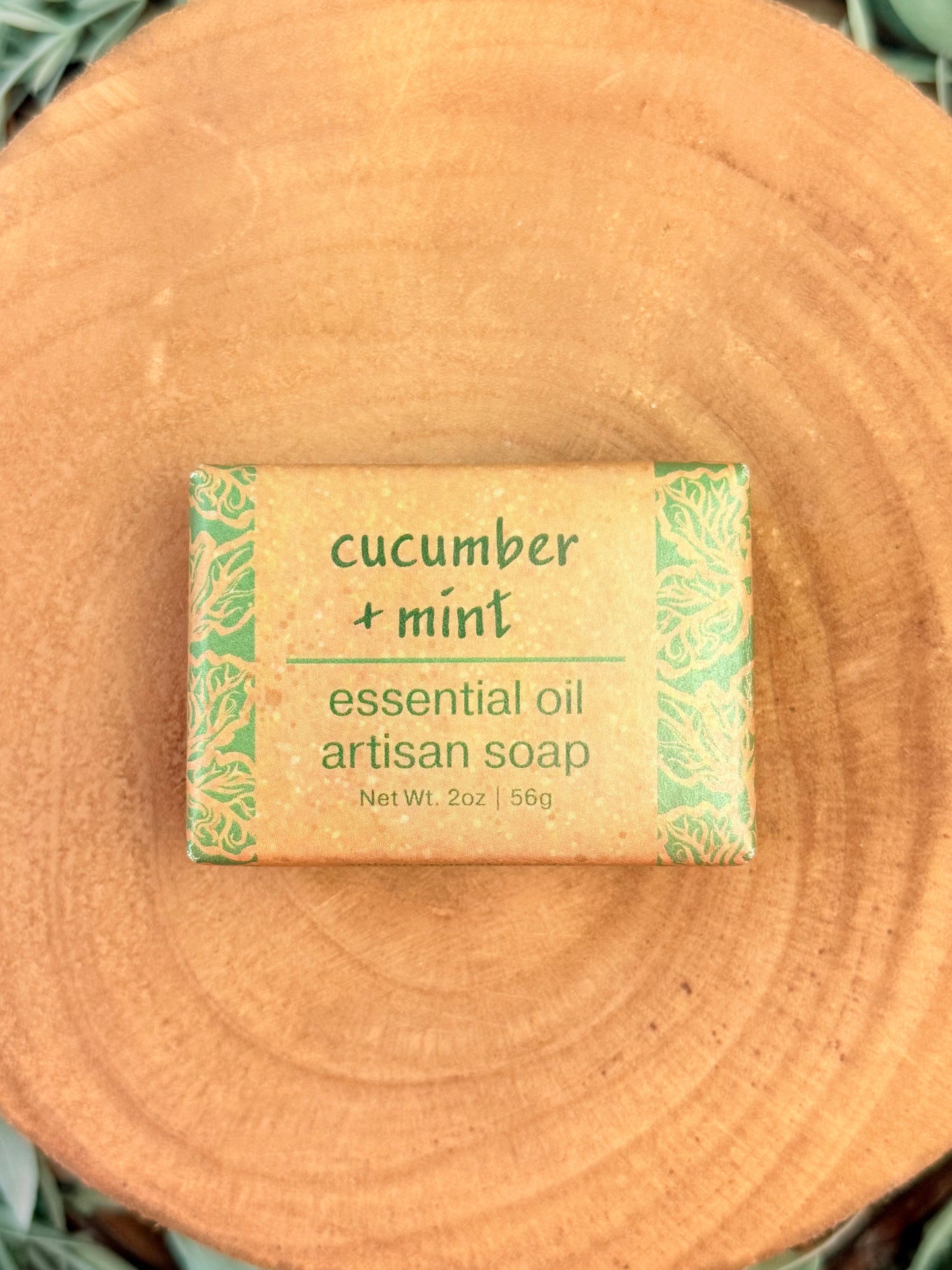 Bar of soap labeled 'cucumber + mint' on a wooden surface with greenery.