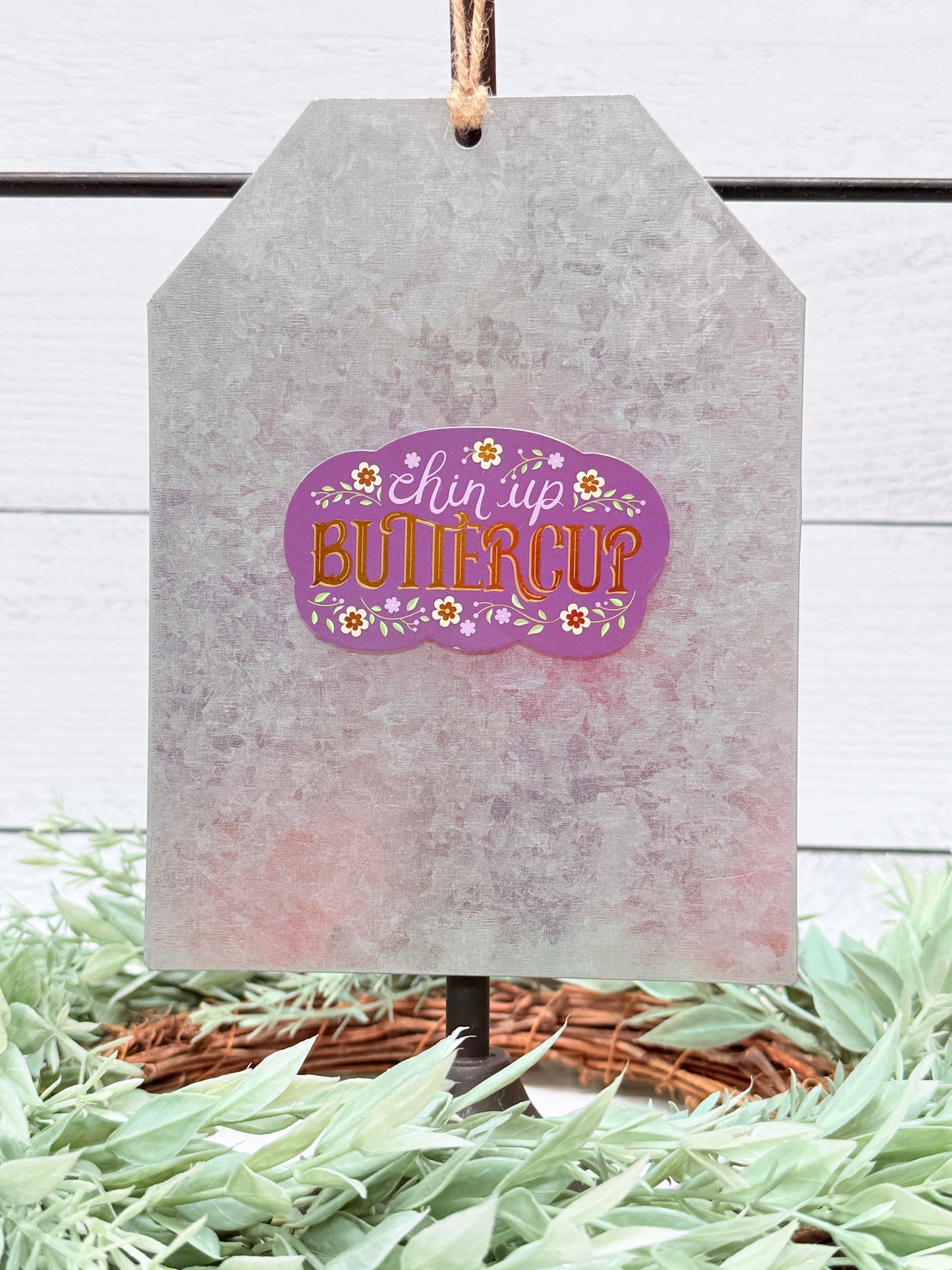 Decorative tag with 'Bless Up Buttercup' text on a white wooden background with greenery.