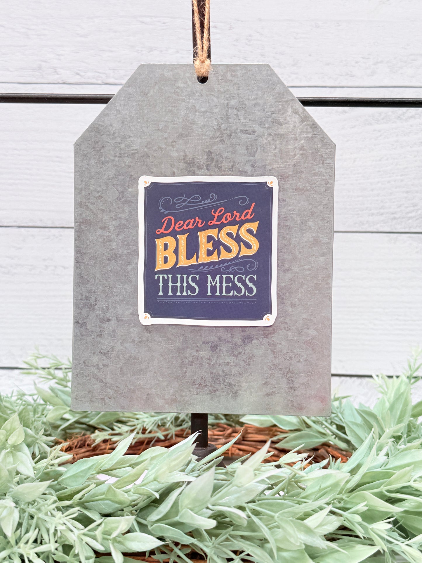 Decorative sign with 'Dear Lord Bless This Mess' text on a wooden background with greenery.