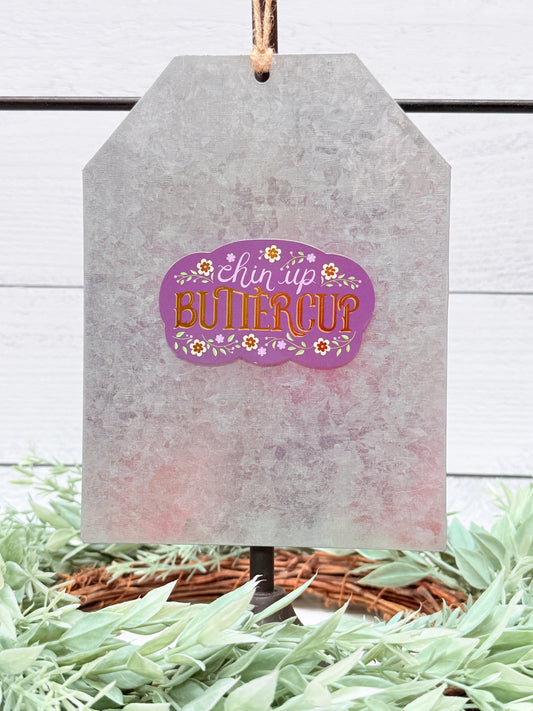 Decorative tag with 'Bless Up Buttercup' text on a white wooden background with greenery.